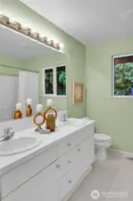 Upper-level guest bathroom. Paneled Quaker door. Tiled double-sink vanity with shiny chrome fixtures, paneled white cabinet doors, shiny-chrome hardware. Mansfield® watercloset. South-facing window with window screen. Fiberglass bathtub with safety hand bar, and shower. Linen closet with coated ventilated wire shelving. Vinyl floor.