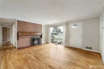Looking back at hardwood floors leading down hallway to the left, brick fireplace, large front window and front door on the right.