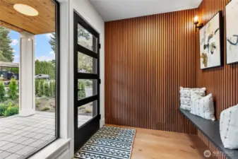 This modern entryway blends warmth and sophistication with vertical wooden slats, abstract art, and a sleek black bench accented by geometric pillows. Natural light pours through the glass-paneled door, framing a tranquil view of the garden and patio beyond.