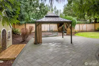 Covered gazebo lounge anchors the paver patio, offering a comfortable all season hangout space surrounded by greenery and garden beds.