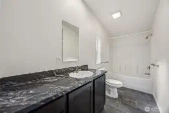 This en suite bathroom understands balance. The long vanity offers real counter space, paired with a dark shaker cabinet that feels modern and substantial. The marble-look Formica countertop is durable, forgiving, and refreshingly low stress. Matching brushed nickel hardware ties the room together, while the privacy-glass window adds daylight and airflow without sacrificing discretion. Stone-style vinyl flooring finishes the space with durability and polish.