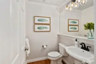 Tucked just off the main hallway between the kitchen and dining room, this elegant powder room features a classic pedestal sink, new faucet, new toilet, updated vanity lighting, and a large wall-to-wall mirror. Warm hardwood floors add a touch of charm to this convenient main-floor space.