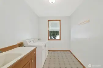 Spacious laundry room with a utility sink, washer and dryer, and ample natural light from the window. Includes wall-mounted coat pegs and generous floor space, ideal for folding or storage solutions.