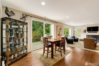 The open dining and living area flows effortlessly to the outdoor deck through large sliding doors, creating a seamless indoor-outdoor living experience. Natural light fills the space, highlighting the beautiful hardwood floors and peaceful, tree-lined backdrop.