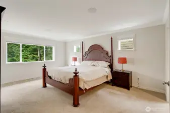 The spacious primary suite offers a peaceful retreat with a wall of windows overlooking lush evergreens. Neutral tones, soft carpeting, and abundant natural light create a calm and private sanctuary for rest and relaxation.