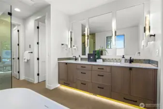 Long double sink vanity, motion-activated toe-kick lighting, and decorative wall sconces. Drawers and cabinets provide ample storage.