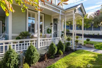 A charming, manicured front garden framed by a white picket fence, mature shade tree, and the home’s picturesque Victorian porch with detailed spindlework.