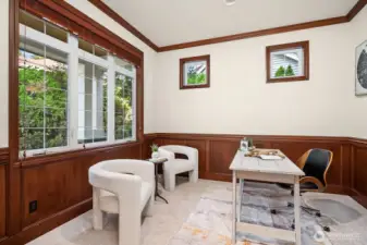 The dedicated home office offers a quiet and functional workspace, featuring rich wood wainscoting, large windows overlooking lush greenery, and a bright, airy atmosphere that inspires productivity. This thoughtfully designed space provides the perfect balance of privacy and natural light for working from home in comfort and style.