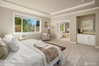 This angle of the expansive primary suite highlights the generous proportions of the space, with large windows flooding the room with natural light and offering tranquil views of the surrounding landscape. A glimpse of the dual walk-in closets beyond underscores the thoughtful functionality that makes this retreat as practical as it is luxurious.