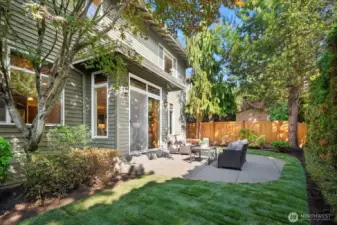 This wonderful full view of the private backyard captures the perfect balance of hardscape and lush landscaping, with a spacious patio flowing into a manicured lawn area surrounded by mature trees and privacy fencing. The home's charming craftsman exterior and abundance of large windows are beautifully showcased from this angle, highlighting the seamless connection between the interior living spaces and this exceptional outdoor retreat.