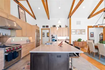 View 2 of the Living Room and Dining Room with soaring ceilings, exposed wood beams, wide plank wood floors, stone fireplace and glass sliding doors leading to the outdoor living area.