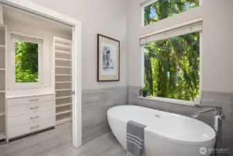 Freestanding soaking tub positioned beneath a large picture window, with adjacent walk-in closet and serene treed outlook.