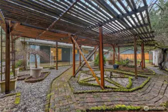 A wide view of the sprawling wood pergola showcases the scale of this outdoor space, with moss-edged brick pathways, gravel beds, and a direct sightline back to the covered patio and home.