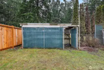 A fenced, covered structure with a corrugated metal roof and concrete base sits along the back fence line, offering flexible use as secure storage, a hobby space, or specialty outdoor enclosure.