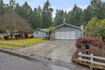 A second street-facing angle highlights the home's wide concrete driveway, two-car garage, and well-established front landscaping, all set against a dramatic backdrop of tall PNW conifers.