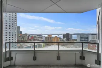 Private Seattle condo balcony overlooking surrounding city buildings with expansive Puget Sound water views and open sky beyond the urban skyline.