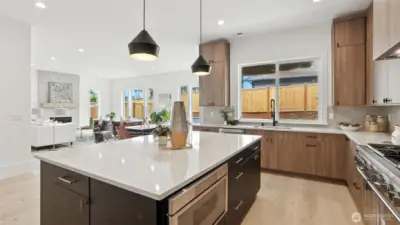 A striking centerpiece for daily life and entertaining alike. The chef’s kitchen pairs rich wood cabinetry with sleek black-and-brass accents, grounded by an oversized quartz island that doubles as a prep station and social hub. Pendant lighting adds modern warmth, while full-height windows draw natural light across the open-concept layout. The seamless flow into the great room highlights the home’s sense of space and connection — perfect for casual gatherings or elegant evenings by the fire.