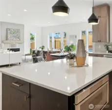 A striking centerpiece for daily life and entertaining alike. The chef’s kitchen pairs rich wood cabinetry with sleek black-and-brass accents, grounded by an oversized quartz island that doubles as a prep station and social hub. Pendant lighting adds modern warmth, while full-height windows draw natural light across the open-concept layout. The seamless flow into the great room highlights the home’s sense of space and connection — perfect for casual gatherings or elegant evenings by the fire.