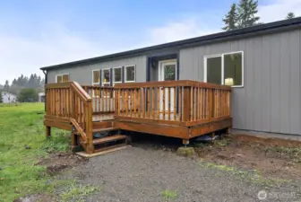 A spacious wood deck with railing leads to the front entry, offering a welcoming arrival with open green space views and mature trees in the background.