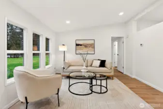 The sunlit living room offers large windows with green views, clean white walls, recessed lighting, and an easy flow to the private bedroom wing.