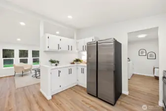 Kitchen peninsula with white cabinetry and stainless refrigerator, open to the living room with views to the yard and a doorway leading to the utility/laundry room.