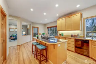 The spacious kitchen is designed for both function and style, featuring an expansive island with a gas cooktop and bar seating. Natural wood cabinetry offers ample storage, while large windows flood the space with natural light. The refinished hardwood floors enhance the warm and inviting ambiance.