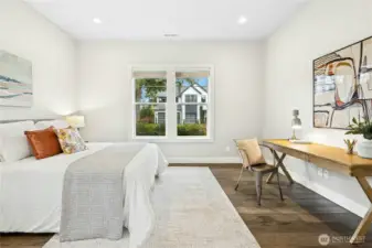 An alternate view of the main-level guest suite highlights its spacious layout and flexible design, easily accommodating both sleeping and workspace needs. Large windows frame neighborhood greenery while maintaining a bright, airy ambiance throughout the room.