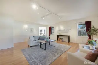 Light-filled living room with hardwoods, updated track lighting, and wall space for art—three picture windows overlook mature landscaping and fenced yard.