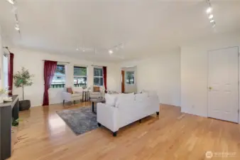 Light-filled living room with hardwoods, updated track lighting, and wall space for art—three picture windows overlook mature landscaping and fenced yard.