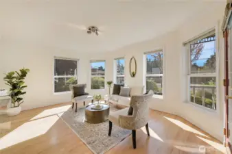 Bright bayed sitting room with wraparound windows, warm wood floors, and peaceful front-garden views—ideal for morning coffee, reading, or intimate conversations.