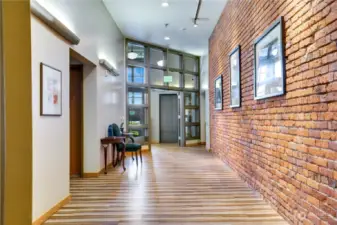 Entering the secure lobby, you are greeted with the essential elements of industrial interior design —exposed brick walls, metal accents, weathered wood, and a neutral color palette. Notice the convenient elevator just ahead! Let's go upstairs!