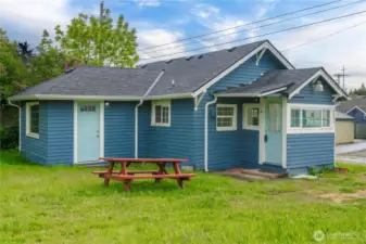 This closer look highlights the charming blue siding, crisp white trim, and two separate entry doors—one into the cozy living room and another through the light-filled laundry/mudroom. The yard is flat and grassy with space for outdoor dining, gardening, or a potential patio upgrade, making this a versatile bonus for buyers looking for low-maintenance outdoor space.
