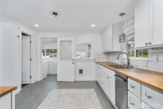 Enjoy a stylish, functional layout featuring butcher block countertops, sleek white cabinetry, and a beautiful tile backsplash. This view also highlights direct access to the bright mudroom/laundry area—perfect for keeping everyday messes out of sight and adding extra utility to this updated home.