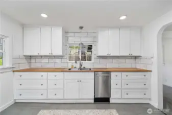 This crisp white kitchen blends charm and function with shaker-style cabinetry, warm butcher block counters, and a marble-look tile backsplash that adds a touch of elegance. A window above the sink brings in natural light, while the custom tile inlay underfoot defines the space with personality. Despite its compact footprint, there’s no shortage of necessities—including a sleek, space-saving dishwasher and plenty of cabinet storage for all your kitchen must-haves.