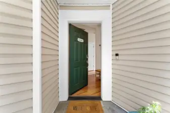A charming covered entry welcomes you with a classic green door, warm wood floors, and a bright, inviting interior just beyond.