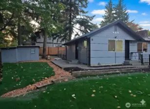 View from the front corner showing both entrances — the main door and the laundry patio access — along with the side yard and storage shed. A clean, functional layout surrounded by mature trees and fresh landscaping.