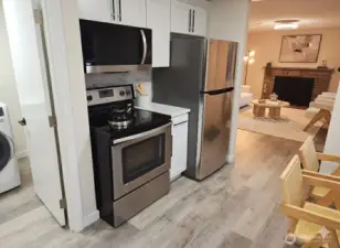 View from the kitchen toward the living room and laundry area — showcasing stainless steel appliances, modern finishes, and a seamless open flow between the main spaces.
