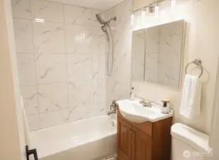 Updated full bathroom with an alcove bathtub and a marble-look tiled shower surround, brushed-nickel showerhead and tub fixtures, and a compact wood-tone vanity with integrated sink. Mirrored medicine cabinet and modern vanity lighting provide bright, even illumination. Neutral walls, clean trim and waterproof laminate flooring complete the space — a fresh, move-in ready bath with contemporary finishes.