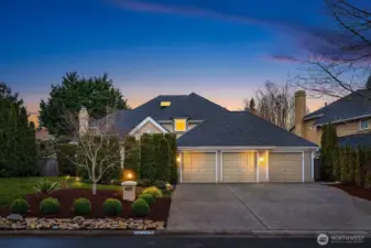 Stunning twilight curb appeal showcases this impeccable Buchan home in the coveted Trossachs community, featuring a three-car garage, mature landscaping, and warm exterior lighting set against a dramatic Pacific Northwest sunset sky.