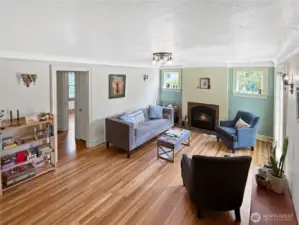 Step into timeless character with this inviting living room, where coved ceilings, original hardwood floors, and vintage sconces blend effortlessly with modern updates. The wood-burning fireplace offers a cozy focal point, flanked by charming cottage-style windows that bathe the space in natural light. Built in an era when craftsmanship mattered, this room is a warm reminder that cool never goes out of style.