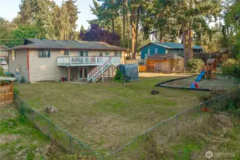 View from the back fence highlights the home’s full deck, lower patio, and large, fully fenced yard — ideal for pets, play, or future landscaping ideas. Mature trees and established greenery add a peaceful Pacific Northwest feel.