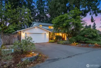 Twilight exterior of a two-story home in Puyallup featuring a two-car garage, landscaped front yard, mature evergreen trees, and warm porch lighting that highlights the welcoming front entry.