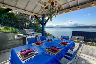 Beneath the shelter of a flower-strung pavilion, the upland terrace transforms into a dreamscape for alfresco dining. A long table dressed in vivid blue stretches toward the horizon, set for a feast with friends. Nearby, a gleaming grill stands ready, framed by a chorus of tulips and hyacinths. As the sun drifts lower over Browns Point and the Sound shimmers below, this space whispers its promise: come for the view, stay for the moment.
