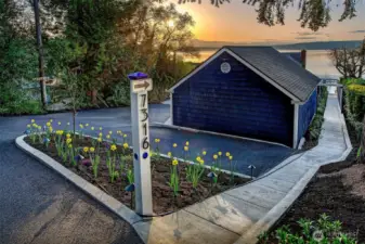 At the edge of a quiet lane, where the trees part just enough for the light to spill through, a sculptural signpost guides your arrival—7316 carved cleanly into its face, flanked by art-glass inlays and a gentle curve of daffodils rising from fresh soil.    Here, the 520-square-foot Coach House stands at the top of the bluff—its midnight-blue shingles catching the last golden light of day. Inside, a freshly finished interior awaits: ample shop space, upgraded electrical service, and secure storage above, accessed by ladder to a hidden loft. It’s a place where tools are kept, projects begin, and the story of the shoreline unfolds from a quiet remove.    A smooth blacktop turnaround eases the transition from road to retreat. And from the Coach House door, a ribbon of poured concrete walkway unfurls, leading you through layers of cultivated beauty, down toward the salt air and shimmer of Puget Sound.