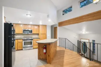 Open-concept kitchen featuring warm wood cabinetry, stainless steel appliances, and a central island with stone countertop. Vaulted ceilings, hardwood floors, and clerestory windows provide abundant natural light and an airy feel, with the kitchen flowing seamlessly into the main living area and stairwell—ideal for everyday living and entertaining.