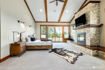 A primary bedroom designed for comfort and elegance, featuring a soaring vaulted ceiling that adds to its grandeur, timber accents highlighting the architectural lines, a gas fireplace with a stone surround and hearth, and direct access to the exterior deck extending the luxury of the bedroom into the great outdoors.