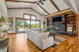 Warm and welcoming great room adorned with a cozy gas fireplace, custom-built wood shelving, rustic stone, soaring vaulted ceilings, and charming timber beam accents.