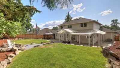 Full view of the wide-open green lawn and rock-covered cement area, highlighting the large covered spaces, perfect for serene and peaceful relaxation. Surrounding dirt areas provide excellent opportunities for gardening and planting