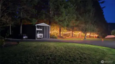 Looking at across the paved, circular and power gated driveway from the home is the 40' x18' RV garage. Fully enclosed on 3 sides for the RV it could easily be fully enclosed and/or doors added. Power, multiple outlets and lighting is in place. Additional parking next to it. The woods to the right are a magical wonderland with separate areas for play or "camping". Even the adults cannot beat the temptation to sit in the swings hanging from the huge fir branches.