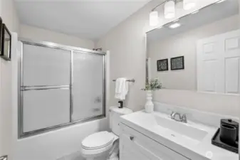Remodeled in 2026, this main floor full bathroom carries a fresh, bright presence, with soft neutral walls and new flooring that give the room a clean, updated look.  The tub and shower are set behind a sliding glass door with textured panels that gently diffuse the light while offering privacy when needed. The white cultured marble vanity provides a smooth, continuous surface beneath the mirror, paired with coordinating brushed nickel faucet and cabinet pulls that create a quiet sense of continuity throughout.  Above the mirror, a brushed nickel vanity light set with frosted glass shades spreads an even, flattering light across the space, keeping the room bright and uplifting glow to the space.